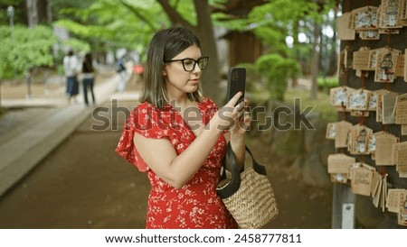 Beautiful hispanic woman in glasses capturing ema prayer boards at japanese gotokuji temple, preserving tradition through a smartphone lens