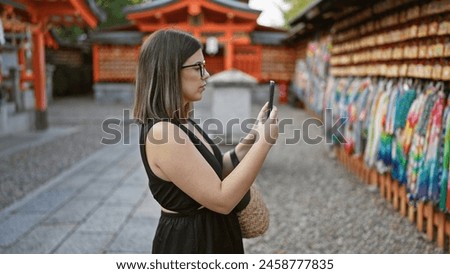 Beautiful young hispanic woman wearing glasses taking pictures to japanese ema wooden boards at fushimi inari-taisha