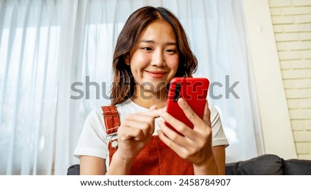 Cheerful young asian woman using smartphone while sitting on couch. Smiling woman using app on cellphone at home. Beautiful girl relaxing while chatting on mobile phone