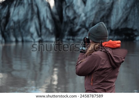 Blonde tourist takes a photo on the island. Photographer with analogue camera. Explorer, hiker, northern lands, ice, distant places. Cold, green hat on head.