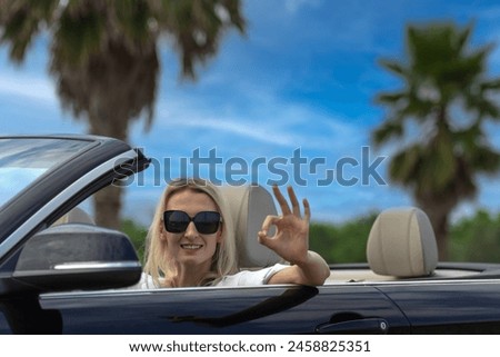 Portrait of a beautiful young woman in a white shirt and sunglasses smiling at the wheel of a convertible with an open roof showing an OK sign against a background of blue sky and palm trees.