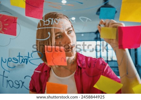 Closeup of young beautiful caucasian business leader presents marketing idea while writing marketing idea on glass board with mind map and colorful sticky notes. Portrait. Brainstorm. Immaculate. Royalty-Free Stock Photo #2458844925
