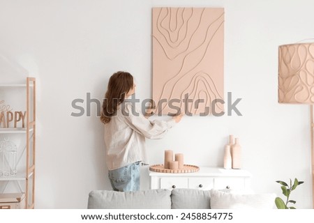 Young pretty woman hanging picture on white wall in room