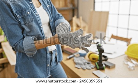 A young woman prepares for carpentry work, donning gloves in a sunlit workshop. Royalty-Free Stock Photo #2458859551