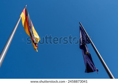 Two flags fly high in the sky, one of which is German. Another flag is the EU flag.