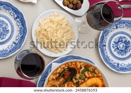 A dish of seafood feijoada with rice, garnished with shrimp and clams, sits on a restaurant table. Alongside are Alentejo cheese and a glass of red wine, evoking a warm Portuguese dining experience.