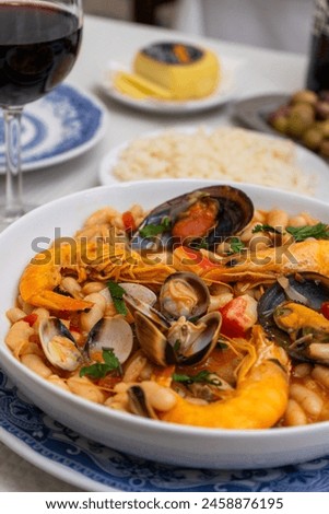 A dish of seafood feijoada with rice, garnished with shrimp and clams, sits on a restaurant table. Alongside are Alentejo cheese and a glass of red wine, evoking a warm Portuguese dining experience.