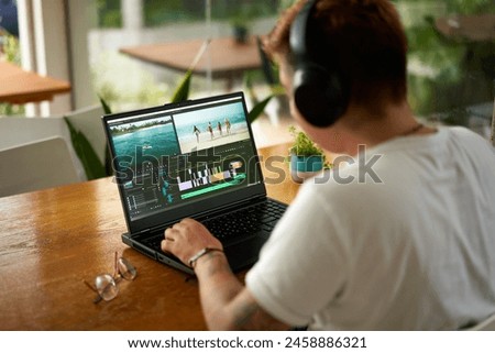 Professional video editor works on laptop with editing software, headphones on. Editing beach scene timeline, focus on creative process in home office. Casual work environment, multimedia project.