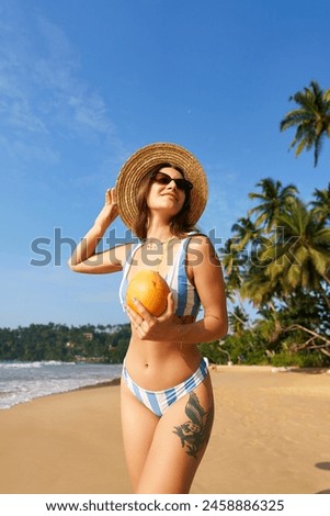 Tanned woman in stylish bikini holds fresh coconut on sunny tropical beach. Fit body, summer fashion, enjoying vacation. Clear blue sky, palm trees provide exotic backdrop. Leisure, travel, wellness.
