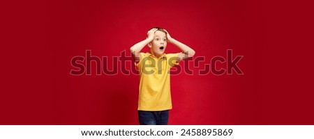 Banner. Surprised, shocked little boy put hands on head of excitement against red studio background with negative space. Concept of human emotions, childhood, education, fashion and style. Ad