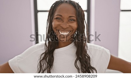A cheerful young woman with curly hair in a white shirt smiling broadly in a brightly lit modern interior. Royalty-Free Stock Photo #2458908209