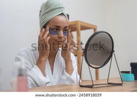 Bathrobe beauty: A lady, in close-up, applies eye patches, gazing at her reflection while perfecting her anti-aging skincare during a home spa day. 