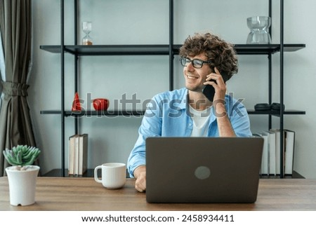 Caucasian young businessman talking on phone while working from home. Attractive handsome freelancer guy sitting on table and talking on smartphone to communicate with coworker in living room at house