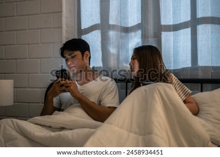 Asian man secretly chatting to mistress while sit on bed with girlfriend. Attractive woman try to look at disloyal husband using phone communicate with paramour lover during night bedtime in bedroom. Royalty-Free Stock Photo #2458934451