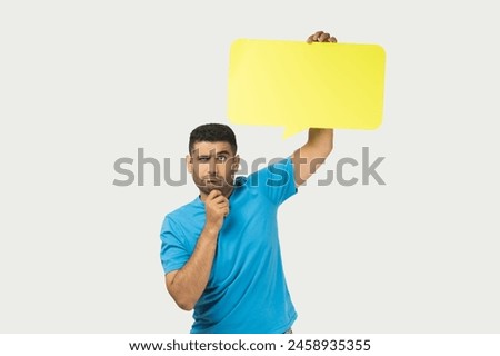 Portrait of thoughtful pensive unshaven man wearing blue T- shirt standing keeps hand on chin, holding yellow speech frame, space for words and thoughts. Indoor studio shot isolated on gray background