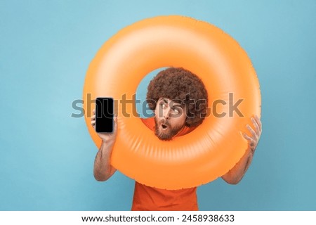 Shocked man with Afro hairstyle holding orange rubber ring, showing phone with blank screen for advertisement, looking at display with open mouth. Indoor studio shot isolated on blue background.