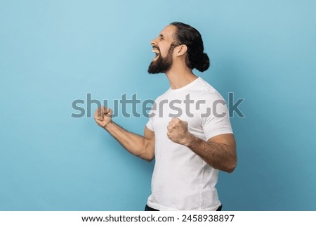 Side view of excited man with beard wearing white T-shirt expressing winning gesture with raised fists and screaming, celebrating victory. Indoor studio shot isolated on blue background. Royalty-Free Stock Photo #2458938897
