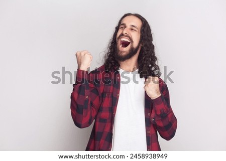 Emotional bearded man raises clenched fists, exclaims with excitement, rejoices sweet success, feels taste of victory, shouts for favorite team. Indoor studio shot isolated on gray background. Royalty-Free Stock Photo #2458938949