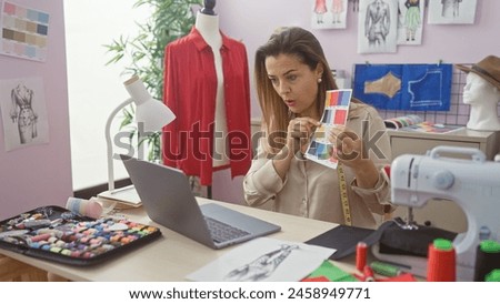 A focused hispanic woman in a tailor shop examines color swatches during a video call, surrounded by design sketches and sewing tools.