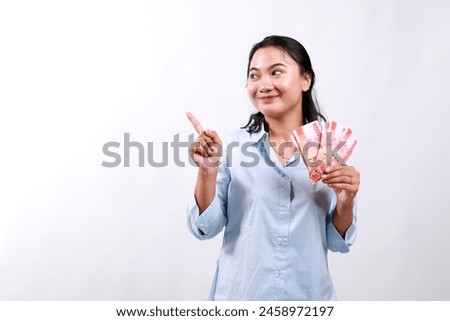 Smiling young modern asian woman, pointing and looking at empty space, holding cash money rupiah, standing over white background