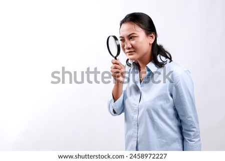 Asian woman detective looking through magnifying glass with intrigued look, found clues, standing over white background. Royalty-Free Stock Photo #2458972227