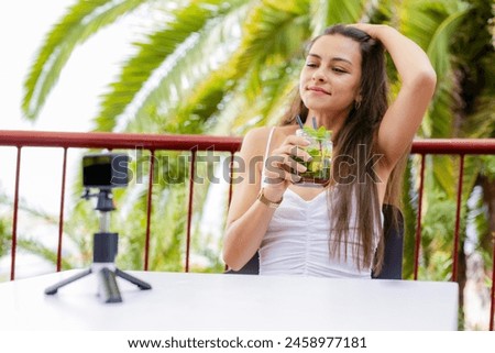 A woman is sitting on a balcony with a camera in front of her. She is smiling and she is enjoying her time. The camera is capturing the moment, and the woman seems to be having a good time