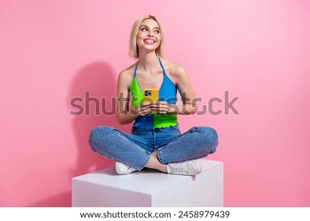 Full size photo of lovely woman wear colorful tank sit on platform look empty space hold smartphone isolated on pink color background
