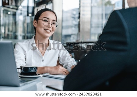 Young man and asian woman discussing work over a cup of coffee sitting at business center cafe, using laptop computer. Businessman and businesswoman partners having a meeting in office lobby. Royalty-Free Stock Photo #2459023421