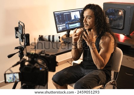 Technology-Driven Video Production. A long-haired content creator immersed in his studio, recording videos with his phone mounted on a tripod. Cameras, lights, and a computer are part of his setup.