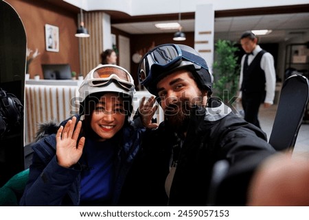 Beautiful couple enjoying winter holiday at ski resort, dressed in winter gear, taking pictures with cell phone. Travelers with skiing and snowboarding equipment capture a photo using mobile device.