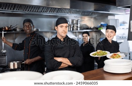Focused experienced man executive chef standing in restaurant kitchen and posing on camera, while his team working Royalty-Free Stock Photo #2459066035