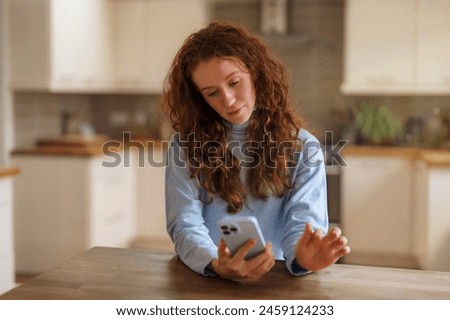 unhappy redhead woman sitting on sofa, using mobile phone, talking, surfing internet, reading, watching videos in kitchen at home. Toned image