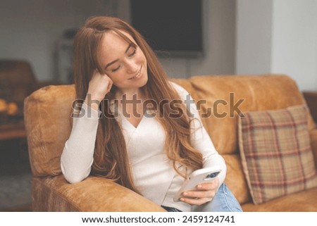 smiling redhead woman sitting on sofa, using mobile phone, talking, surfing internet, reading, watching videos in living room at home. Toned image