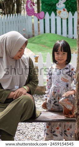 Asian mother in hijab and Asian daughter interacting with a red iguana at the zoo. Royalty-Free Stock Photo #2459154545
