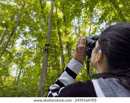 Birdwatching. Photo with a woman filmed from behind while searching and scouting the forest canopy for birds with a binocular.