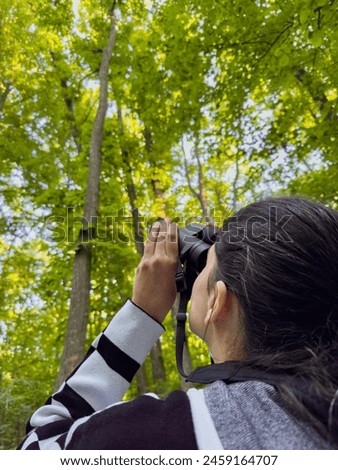 Birdwatching. Vertical Photo with a woman filmed from behind while searching and scouting the forest canopy for birds with a binocular.