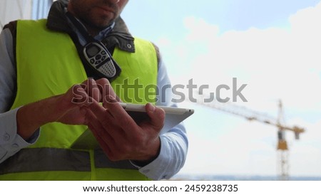 Close up shot of male engineer in uniform standing at construction site and tapping on tablet using gadget outdoor at workplace. Builder typing on device. Building business