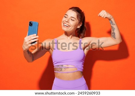 Young fun fitness trainer woman sportsman wear top shorts purple clothes in home gym do selfie shot on mobile cell phone show biceps isolated on plain orange background. Workout sport fit abs concept