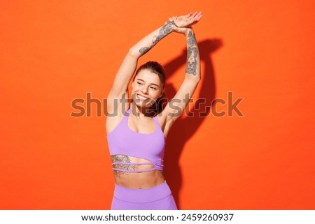 Young fitness trainer sporty woman sportsman wearing purple top clothes spend time in home gym doing stretch exercise rising hands up isolated on plain orange background. Workout sport fit abs concept