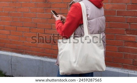 Young woman in red hoodie holding tote canvas blank eco bag and smart phone in hands. street wall background. Female consumer hold white textile shopper. template or place for your design, logo, text