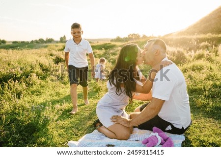 Father kisses mother and little girl, boy having fun on blanket in green grass, nature. Happy young family on vacation together. Portrait of mom embrace dad, son playing with daughter. Holiday picnic