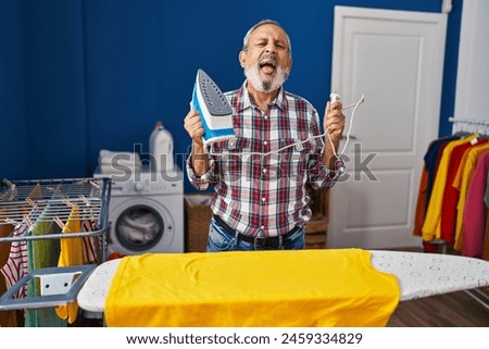 Cheerful senior man having fun in the laundry room, hilariously sticking out tongue while ironing - truly amusing expression of joy!