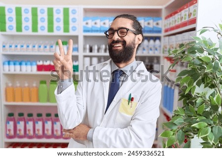 Hispanic man with beard working at pharmacy drugstore smiling with happy face winking at the camera doing victory sign. number two. 