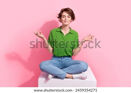 Full size photo of peaceful focused woman dressed green shirt jeans sit on cube in meditation pose isolated on pink color background