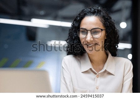 A professional Hispanic businesswoman is happily working on her laptop in a brightly lit office. Her cheerful demeanor radiates positivity and confidence. Royalty-Free Stock Photo #2459415037