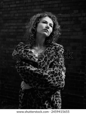 Classic portrait of young woman posing in front of a brick wall. She is wearing a vintage fake fur coat and the soft morning light is lighting her face. Analogue film photography.