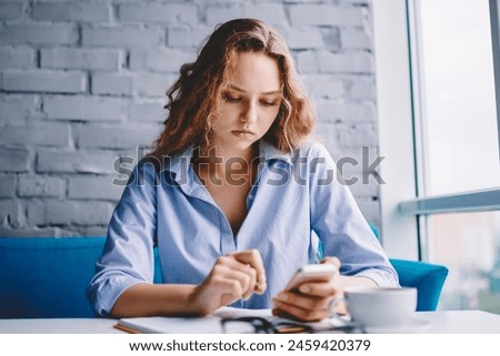 Serious woman with curly hair reading sms message on digital smartphone using free 4g connection in cafeteria, concentrated hipster girl casual dressed watching vied via media application on cellular
