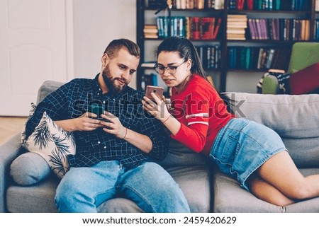 Positive male and female hipster couple viewing common pictures on smartphone sitting on sofa,young marriage share multimedia content via mobile phone and wireless connection at home interior