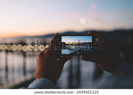 Cropped image of man taking photo in evening dusk of nature environment using modern smartphone camera, back view of hipster guy holding mobile phone making picture of sea water via app in twilight