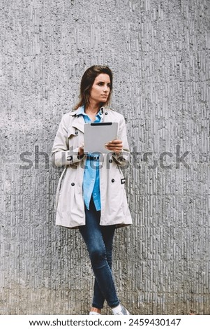 Thoughtful caucasian young woman dressed in casual coat looking away while installing app on modern touch pad with 4G internet.Pondering female blogger with tablet standing on promotional background